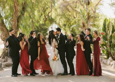 a bride and groom kiss in the middle of a tropical driveway while the groomsmen and bridesmaids cheer