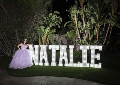 A woman in a purple wedding dress stands in front of several lit up letters that spell out "Natalie"